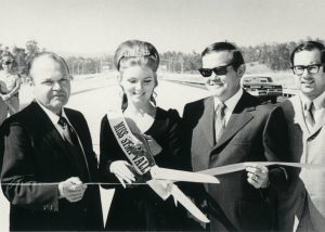 Four people (all of whom are white) posing and smiling for a ceremonial ribbon-cutting ceremony to mark the official opening of the Simi Freeway/SR 118 in 1970. A woman wearing a "Miss Simi Valley" sash stands in the middle of the group holding a large pair of scissors. The group stands in the middle of the freeway with a few cars and people in the background.