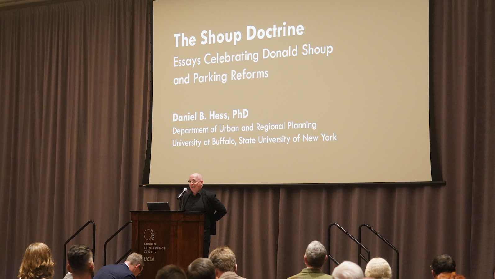 Daniel Hess speaks at a podium in UCLA’s Luskin Conference Center with a presentation slide behind him reading, “The Shoup Doctrine: Essays Celebrating Donald Shoup and Parking Reforms.” The audience is seated in front of him.