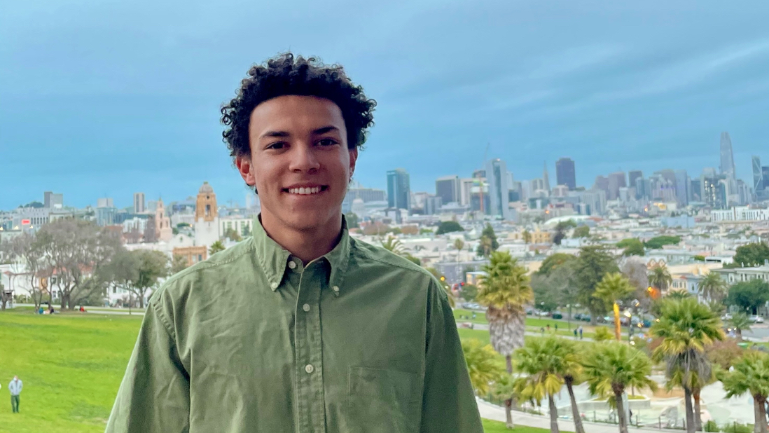 smiling man in green shirt stands in front of a park, palm trees. Behind him further in distance is city skyline